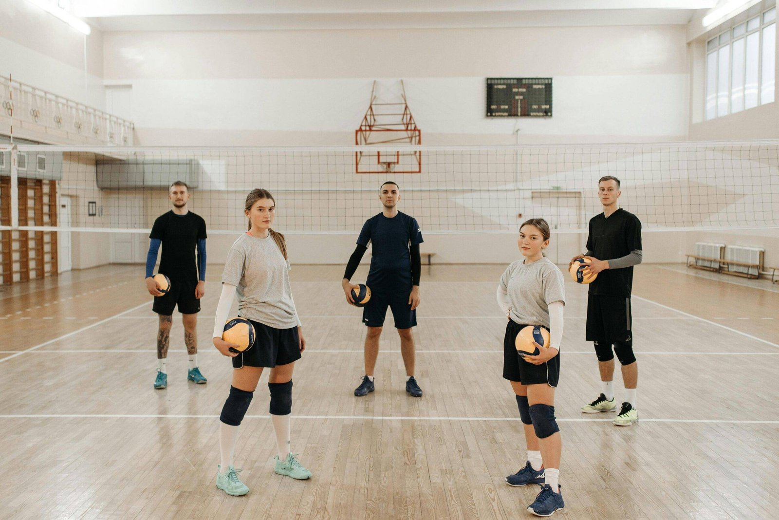 Five volleyball players stand on a beautiful and stylish indoor volleyball court with volleyballs in each hand with the best indoor volleyball shoes.