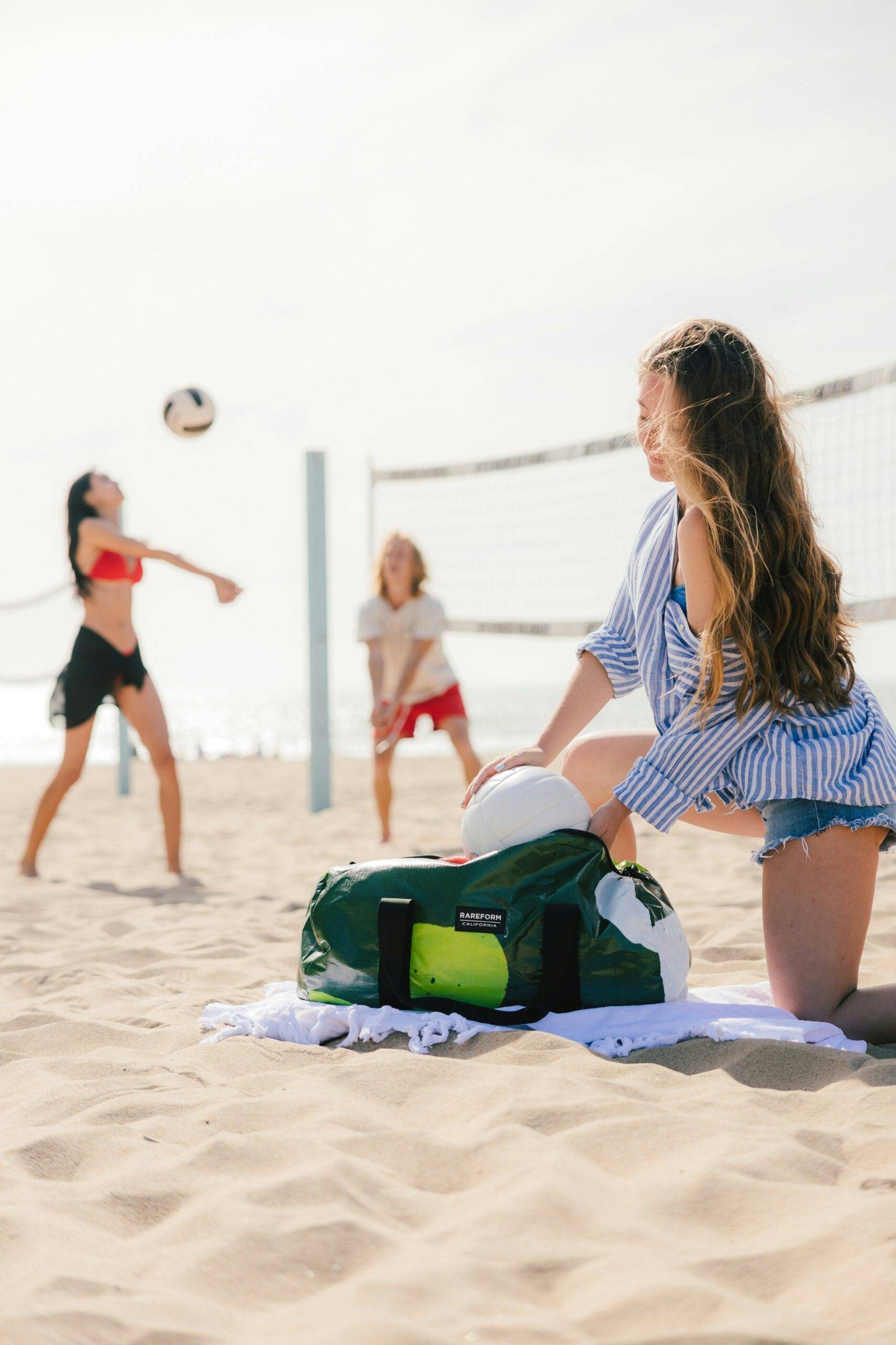 A girl is on a beach volleyball court packing volleyballs and volleyball equipment in a bag while other girls play with volleyballs as practice and wear best beach volleyball shoes