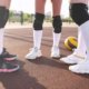 Three players with their best volleyball shoes standing in a outdoor court ahead of a yellow volleyball