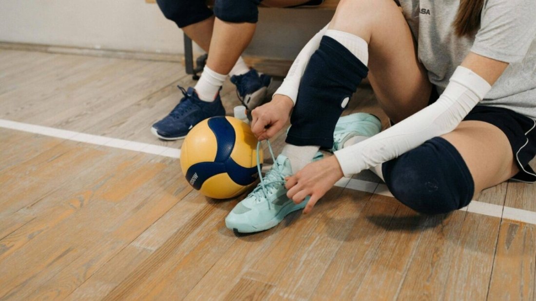 Girl tying her shoe laces with a volleyball by her side, sitting on a gym floor. with comfortable volleyball shoes