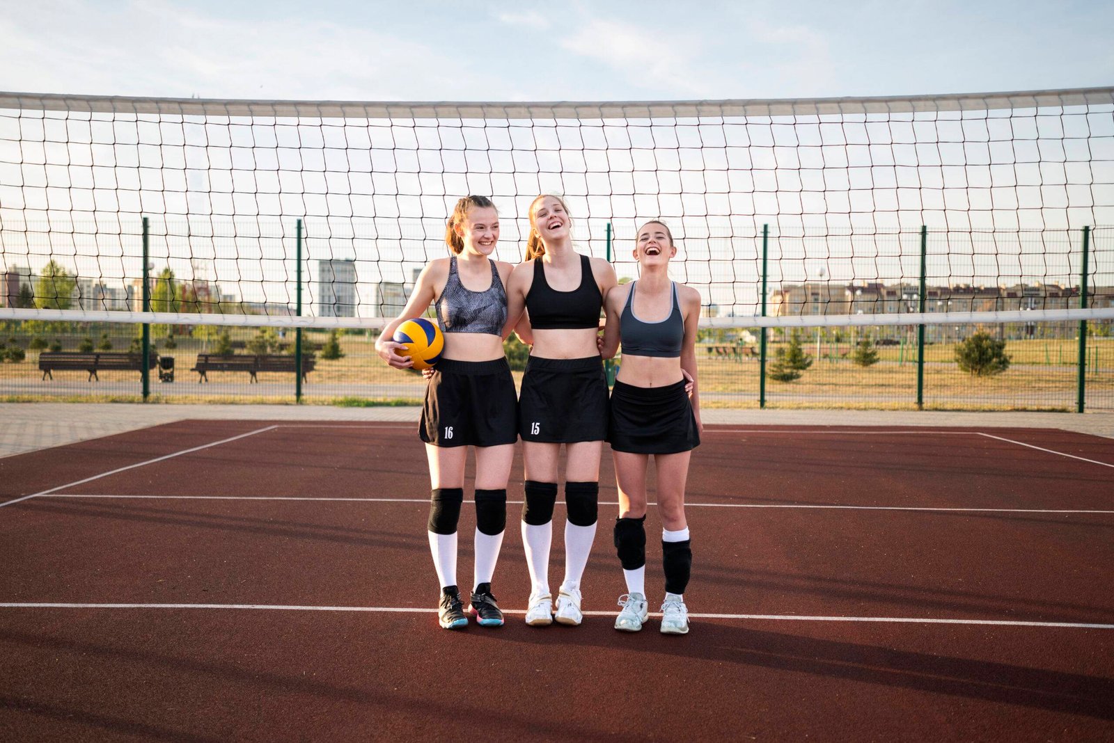 Three girls standing and smiling on an outdoor volleyball court, enjoying the game in durable volleyball shoes.