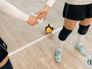 Two women holding hands in indoor volleyball court with a volleyball in background